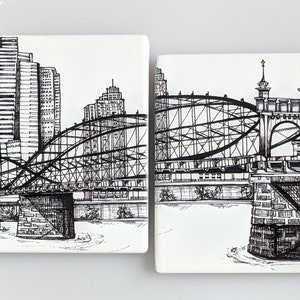 May include: Two black and white ink drawings on ceramic tiles depicting a bridge over a river with a cityscape in the background. The bridge is a steel truss bridge with a curved arch. The cityscape includes tall buildings.