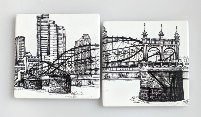 May include: Two black and white ink drawings on ceramic tiles depicting a bridge over a river with a cityscape in the background. The bridge is a steel truss bridge with a curved arch. The cityscape includes tall buildings.
