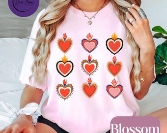 Könnte beinhalten: Ein hellrosa T-Shirt mit neun Herzmotiven mit Flammen. Die Herzen sind rot, rosa und weiß mit dekorativen Bordüren. Das Wort "Blossom" ist in Weiß auf einem lila Rechteck gedruckt.