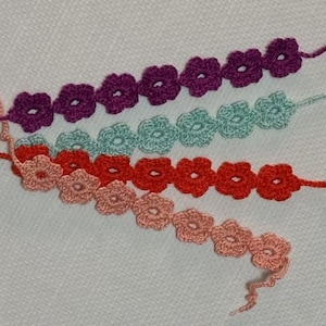 Pode incluir: Quatro fileiras de guirlandas de flores de crochê em tons de roxo, verde-azulado, vermelho-alaranjado e pêssego. Cada guirlanda apresenta múltiplas formas de flores pequenas e detalhadas, conectadas por um fio fino. As guirlandas estão dispostas diagonalmente sobre uma superfície branca.