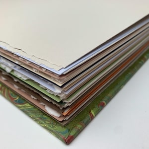 May include: A thick book with a green marbled cover featuring a peacock feather pattern. The pages have rough, deckled edges and are stacked in layers of varying colours including cream, white, light green, brown, and orange, showing the binding.