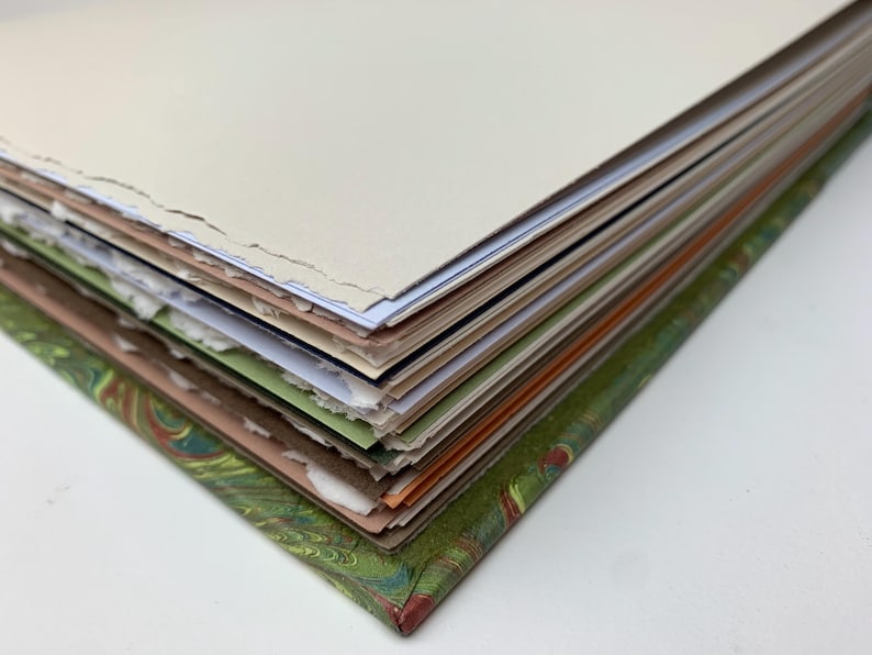May include: A thick book with a green marbled cover featuring a peacock feather pattern. The pages have rough, deckled edges and are stacked in layers of varying colours including cream, white, light green, brown, and orange, showing the binding.
