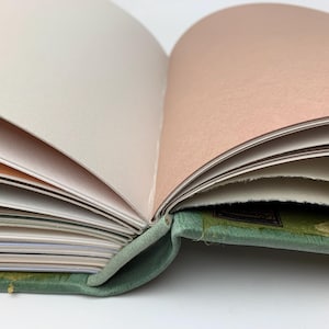 May include: An open book with a sage green fabric cover and a visible spine binding. The interior pages feature a variety of colours, including white, cream, and soft peach, with some pages displaying rough, deckled edges along the side.