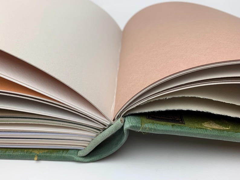 May include: An open book with a sage green fabric cover and a visible spine binding. The interior pages feature a variety of colours, including white, cream, and soft peach, with some pages displaying rough, deckled edges along the side.