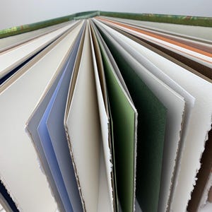May include: A hardback book with a green patterned spine, fanned open to reveal numerous pages of varying colours including white, cream, light blue, sage green, dark green, brown, and orange, featuring textured edges and thick paper stock.