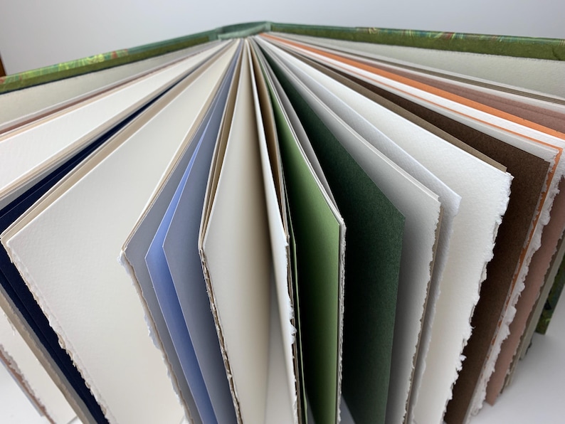 May include: A hardback book with a green patterned spine, fanned open to reveal numerous pages of varying colours including white, cream, light blue, sage green, dark green, brown, and orange, featuring textured edges and thick paper stock.