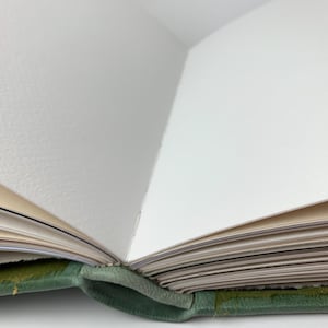 May include: An open book with a sage green leather cover and thick, textured white pages. The binding is visible at the centre fold, showing the stitching that holds the pages together. The paper edges are clean and appear to be high-quality stock.