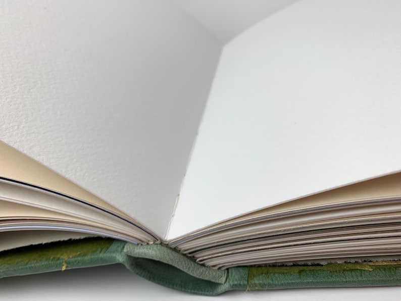 May include: An open book with a sage green leather cover and thick, textured white pages. The binding is visible at the centre fold, showing the stitching that holds the pages together. The paper edges are clean and appear to be high-quality stock.