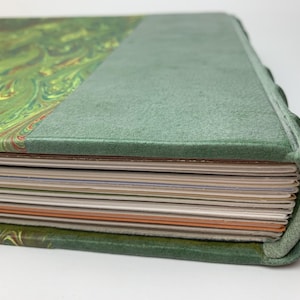 May include: A hardback book with a green marbled paper cover and a solid green spine. The book features a rounded spine edge and a stack of pages with varying colours visible along the fore-edge, including white, cream, and orange paper sheets.