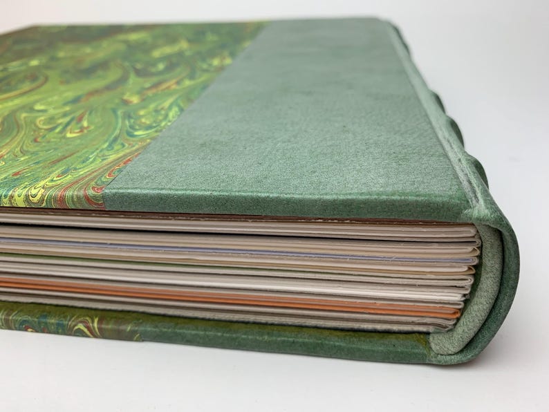 May include: A hardback book with a green marbled paper cover and a solid green spine. The book features a rounded spine edge and a stack of pages with varying colours visible along the fore-edge, including white, cream, and orange paper sheets.