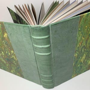 May include: A hardback book with a green suede spine featuring five raised bands. The front and back covers are decorated with a marbled pattern in shades of green, yellow, and brown. The pages are fanned open, revealing white and coloured paper.