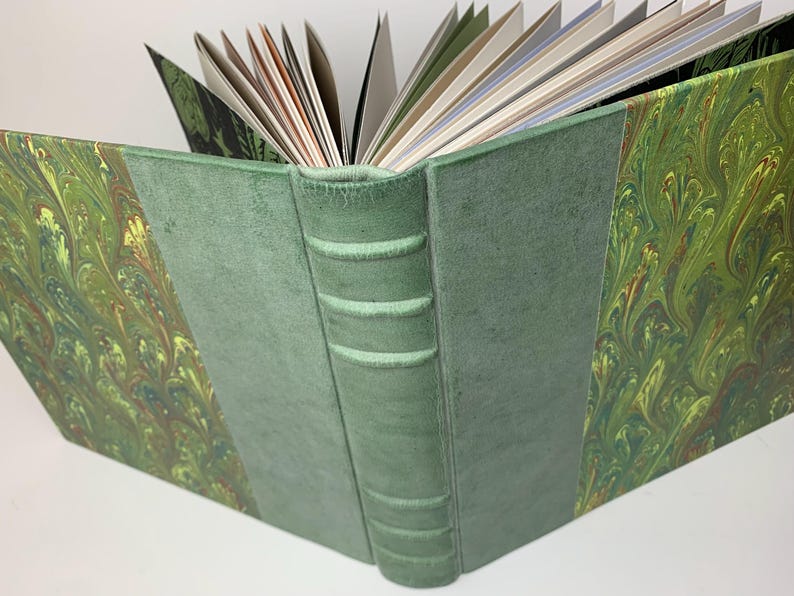 May include: A hardback book with a green suede spine featuring five raised bands. The front and back covers are decorated with a marbled pattern in shades of green, yellow, and brown. The pages are fanned open, revealing white and coloured paper.