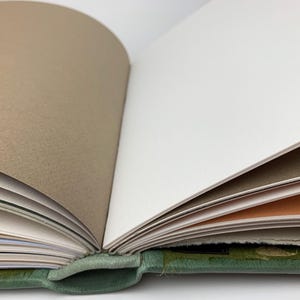 May include: An open book with a green textured cover and multiple pages in alternating colours of white, light brown, and a soft peach tone. The binding is visible at the centre, showing the stack of paper sheets fanned out from the spine.