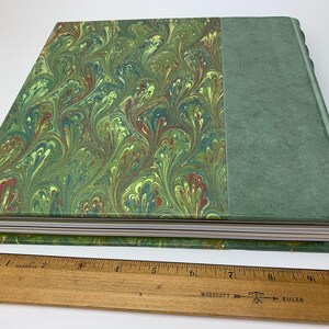 May include: A hardback book with a green marbled paper cover and a solid green spine, placed beside a wooden ruler showing the book is approximately 25.4 cm long. The marbled pattern displays swirling green, yellow, and red colour tones.