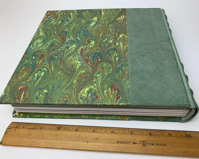 May include: A hardback book with a green marbled paper cover and a solid green spine, placed beside a wooden ruler showing the book is approximately 25.4 cm long. The marbled pattern displays swirling green, yellow, and red colour tones.