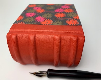 May include: A red, textured book with a floral patterned cover in shades of pink, orange, and brown on a dark grey background. A black fountain pen is in the foreground. The book has a unique, ridged design.