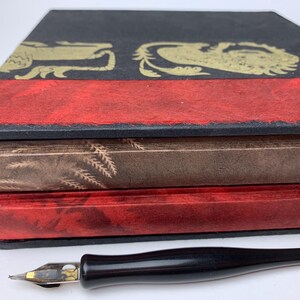 May include: A black box with a gold lion pattern and a red band, containing two stacked books with textured covers in brown and red. A black dip pen with a metal nib is positioned in front of the box on a white surface.