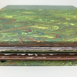May include: A stack of paper sheets featuring a marbled pattern in shades of green, yellow, and brown. The top and bottom sheets have a smooth finish, while the interior pages have rough, deckled edges in white, light green, and tan colours.