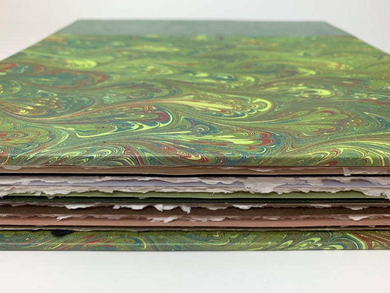May include: A stack of paper sheets featuring a marbled pattern in shades of green, yellow, and brown. The top and bottom sheets have a smooth finish, while the interior pages have rough, deckled edges in white, light green, and tan colours.