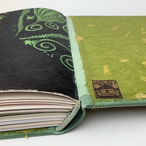 May include: A handcrafted book with a black cover featuring green botanical illustrations and a light green spine. The back cover is textured lime green with gold flecks and a brown label reading slow art studio & bindery.