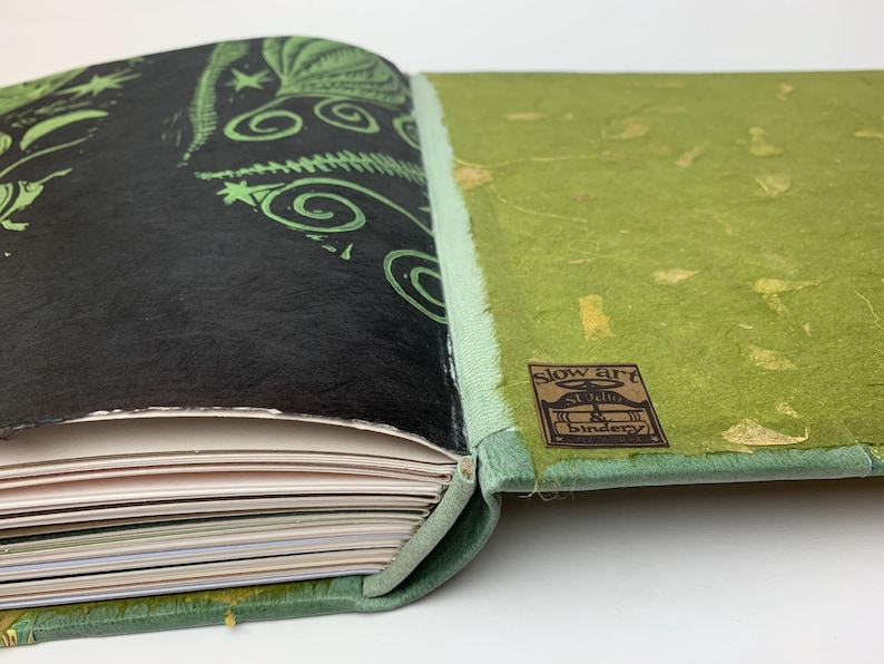 May include: A handcrafted book with a black cover featuring green botanical illustrations and a light green spine. The back cover is textured lime green with gold flecks and a brown label reading slow art studio & bindery.