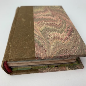 May include: A hardcover book featuring a brown cloth spine and a marbled paper cover with a swirling pattern of pink, green, and tan colors. The book has visible page edges and a red fabric binding detail at the spine, showing a classic design.