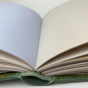 May include: An open book with a green fabric cover and multiple pages. The pages are visible in varying shades of light blue, cream, and white. The binding is visible at the bottom centre, showing the green material connecting the cover to the pages.