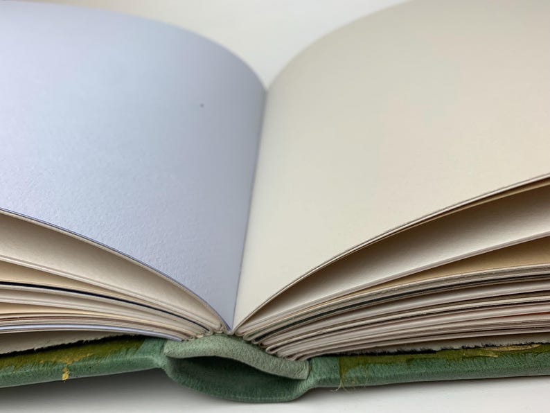 May include: An open book with a green fabric cover and multiple pages. The pages are visible in varying shades of light blue, cream, and white. The binding is visible at the bottom centre, showing the green material connecting the cover to the pages.