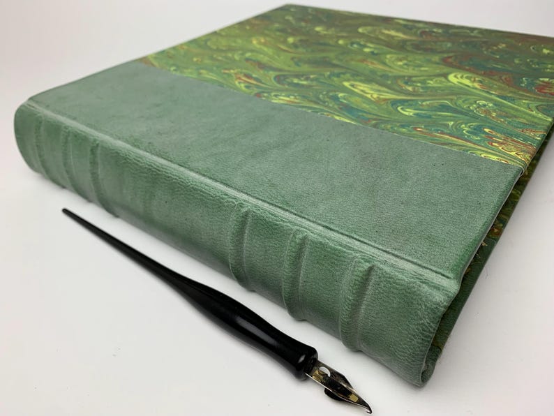 May include: A hardbound book with a sage green textured spine featuring raised bands and a cover with a marbled pattern in shades of green, gold, and brown. A black fountain pen with a silver metal nib is positioned alongside the book.