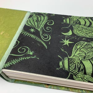 May include: A hardback book with a textured lime green cover and a black endpaper featuring a green block-print design of insects, stars, and a crescent moon. The pages are visible along the edge, and the binding is a light green fabric.
