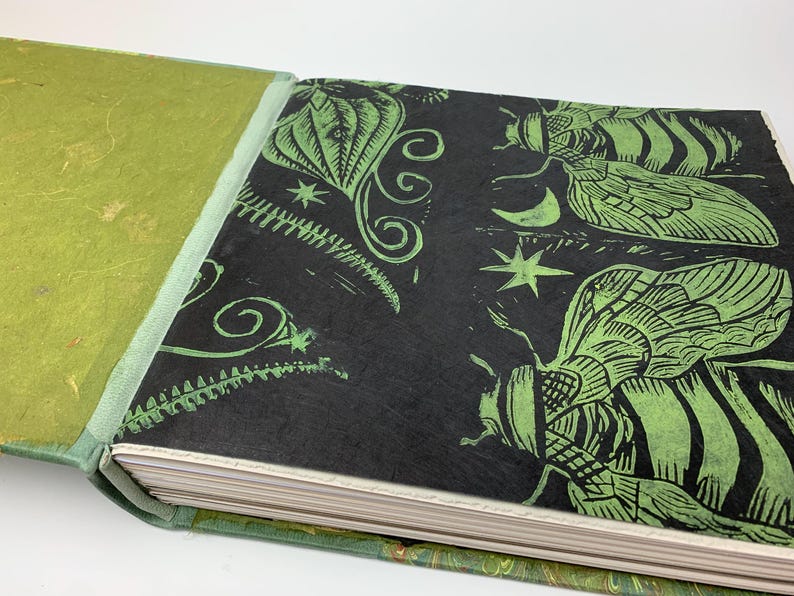 May include: A hardback book with a textured lime green cover and a black endpaper featuring a green block-print design of insects, stars, and a crescent moon. The pages are visible along the edge, and the binding is a light green fabric.