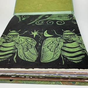 May include: A square book with a black cover featuring a green block-print design of two large insects, a crescent moon, and a star. The book has thick, textured, deckled-edge pages in shades of white, cream, and brown, with a green endpaper visible.