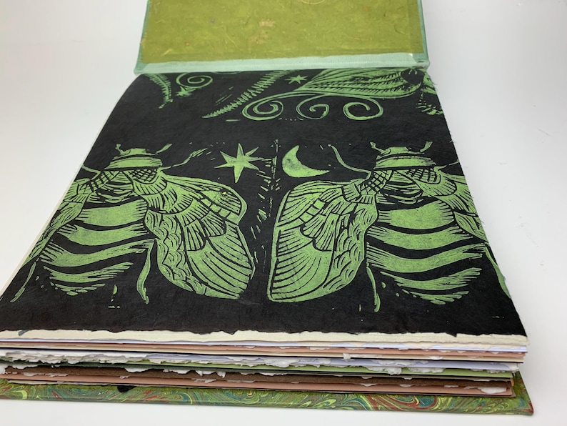 May include: A square book with a black cover featuring a green block-print design of two large insects, a crescent moon, and a star. The book has thick, textured, deckled-edge pages in shades of white, cream, and brown, with a green endpaper visible.