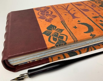 May include: A handcrafted journal with a burgundy leather cover and an orange patterned paper cover featuring floral and celestial designs. A black calligraphy pen lies beside the journal, ready for writing.