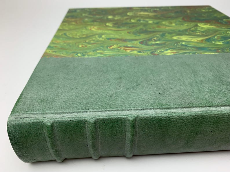 May include: A hardback book featuring a textured, matte sage green spine with four raised bands. The cover displays a marbled pattern in shades of lime green, forest green, and hints of brown, creating a fluid, swirling design across the surface.