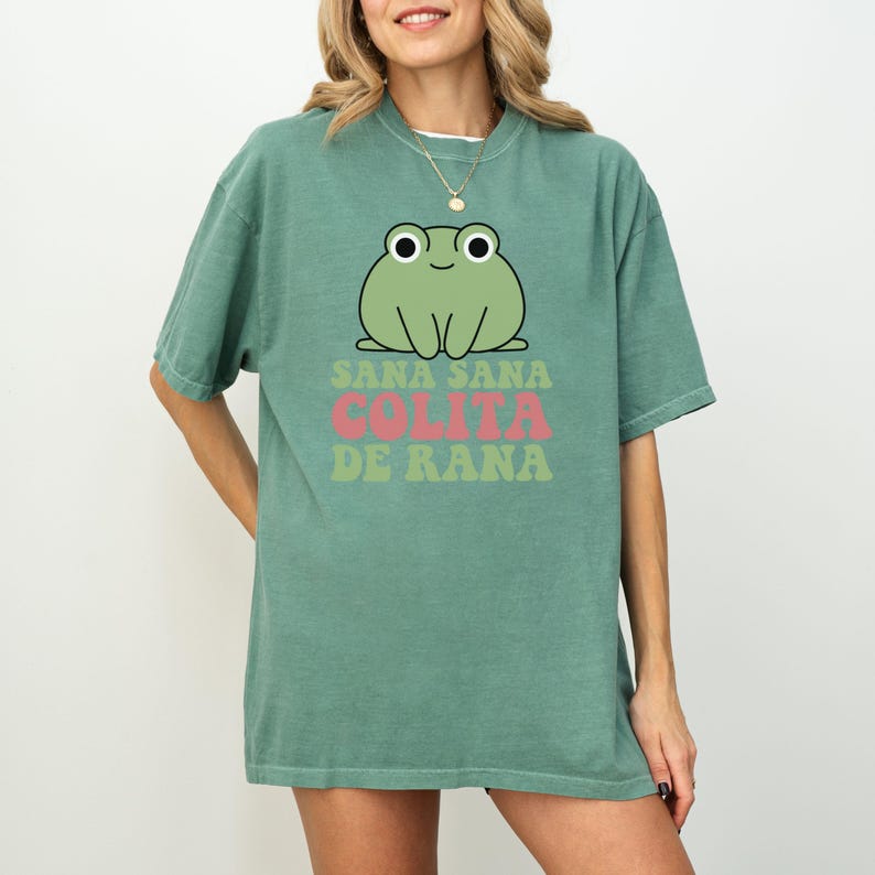 May include: A sage green t-shirt featuring a cartoon frog illustration with the text "Sana Sana Colita de Rana" in pink and green. The frog has large eyes and a smiling expression. The shirt is a relaxed fit.
