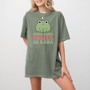 May include: Olive green t-shirt featuring a cartoon frog with the text "Sana Sana Colita De Rana" in pink and green. The frog has large eyes and a smiling expression. The shirt is a relaxed fit.