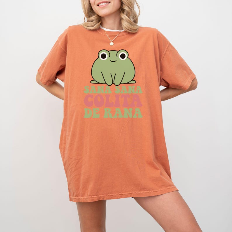 May include: An orange t-shirt featuring a cartoon frog with the text "Sana Sana Colita De Rana" in green and pink. The frog is green with black eyes and is centered above the text. The shirt has short sleeves and a relaxed fit.
