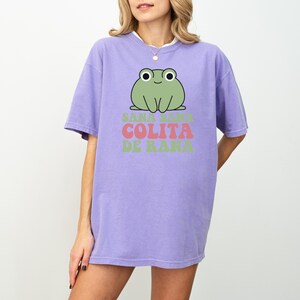 May include: A lilac-colored t-shirt featuring a cartoon frog graphic. The frog is green with white eyes and is above the text "Sana Sana Colita De Rana" in pink and green. The shirt has short sleeves and a relaxed fit.