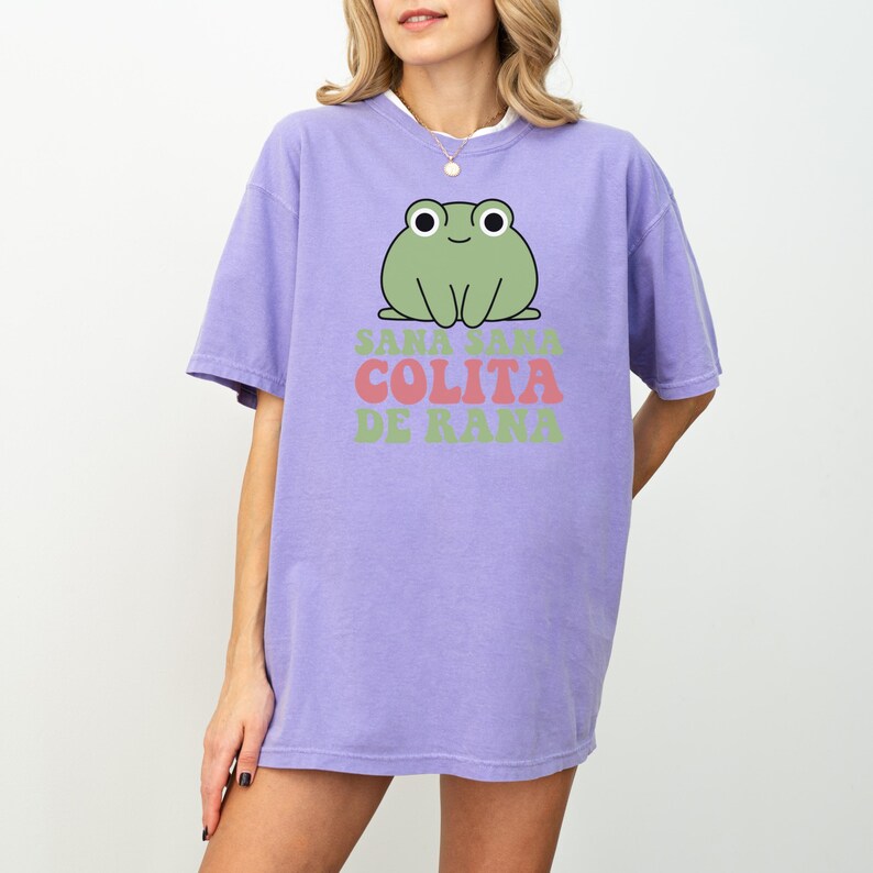 May include: A lilac-colored t-shirt featuring a cartoon frog graphic. The frog is green with white eyes and is above the text "Sana Sana Colita De Rana" in pink and green. The shirt has short sleeves and a relaxed fit.