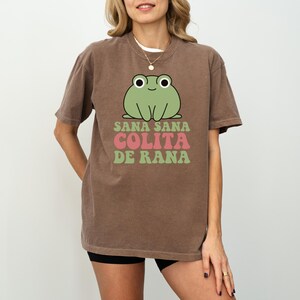 May include: Brown t-shirt featuring a green cartoon frog with the text "Sana Sana Colita de Rana" in pink and green. The shirt has short sleeves and a crew neck.