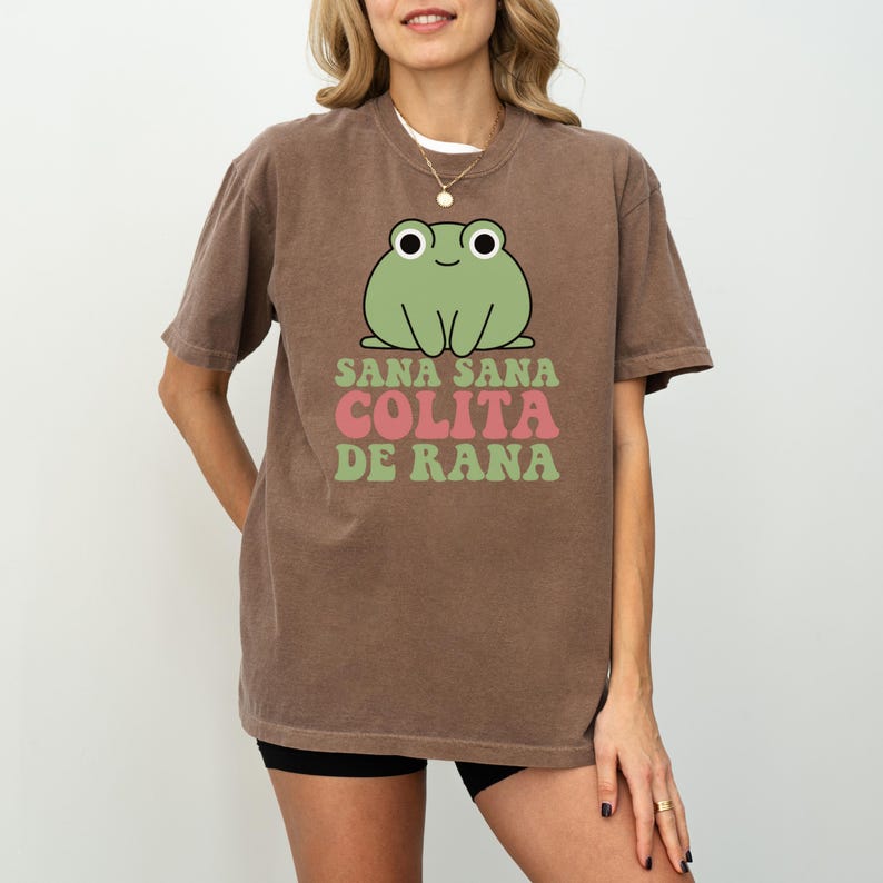 May include: Brown t-shirt featuring a green cartoon frog with the text "Sana Sana Colita de Rana" in pink and green. The shirt has short sleeves and a crew neck.