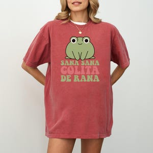 May include: A red-orange t-shirt featuring a cartoon frog with the text "Sana Sana Colita de Rana" in green and pink. The frog is light green with large white eyes and a smiling expression. The shirt has short sleeves.