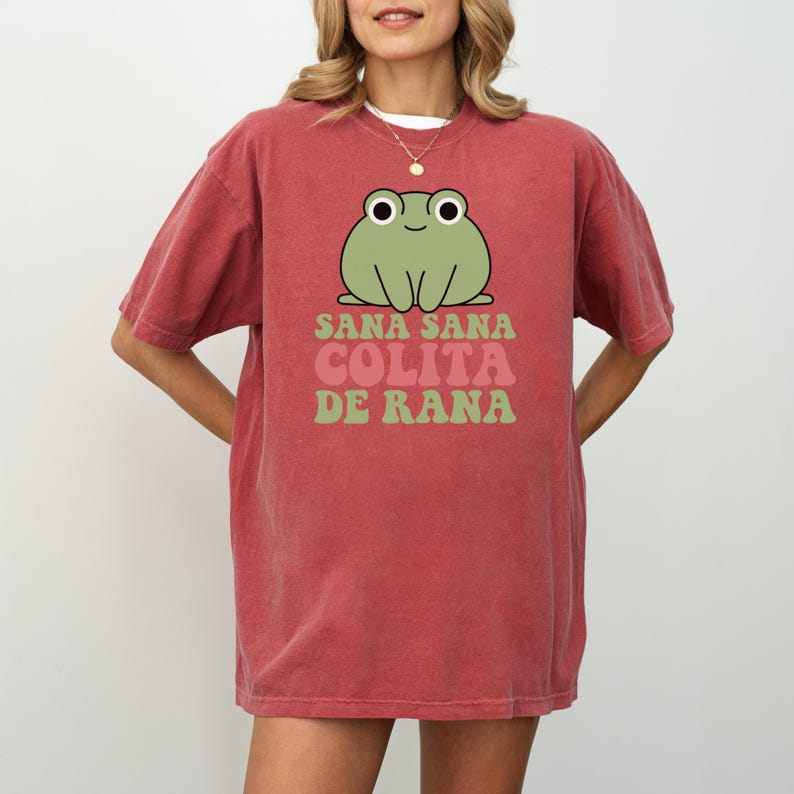 May include: A red-orange t-shirt featuring a cartoon frog with the text "Sana Sana Colita de Rana" in green and pink. The frog is light green with large white eyes and a smiling expression. The shirt has short sleeves.