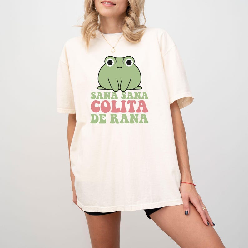 May include: Cream-colored t-shirt featuring a cartoon frog illustration with the text "Sana Sana Colita De Rana" in green and pink. The frog is green with black eyes. The shirt has short sleeves and a relaxed fit.