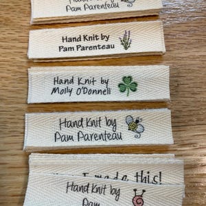 May include: A stack of five white fabric labels with black text that reads "Hand Knit by" followed by a name. Each label has a small graphic image. The images include a bee, lavender, a shamrock, a bee, and a sewing needle and yarn.