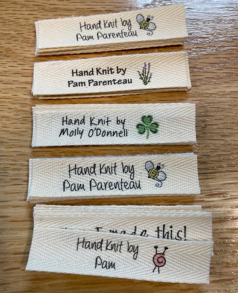 May include: A stack of five white fabric labels with black text that reads "Hand Knit by" followed by a name. Each label has a small graphic image. The images include a bee, lavender, a shamrock, a bee, and a sewing needle and yarn.