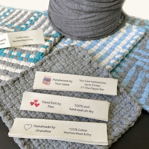 May include: Four white fabric labels with different text and designs. The labels are on a gray woven fabric background. The labels read: "Handwoven by Your name", "Your Care Instructions: Up to 3 Lines 21 Char/Sp per line", "Hand Knit by Pam 100% wool hand wash air dry", and "Handmade by Grandma 100% Cotton Machine Wash & Dry".