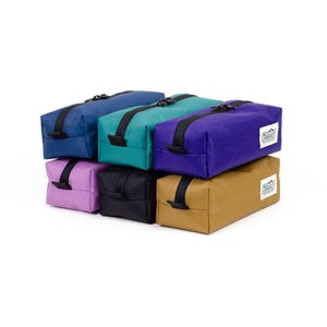 Puede incluir: Un conjunto de estuches rectangulares con cremallera en varios colores: azul marino, turquesa, morado, lila, negro y beige. Cada estuche tiene una cremallera negra y una pequeña etiqueta blanca con un gráfico de montaña.