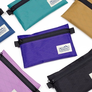 May include: A collection of small, rectangular zippered pouches in various colors, including teal, purple, tan, black, and lavender. Each pouch features a black zipper and a white label with the text "CRAGFAST MADE IN SCOTLAND". The pouches have a diamond pattern.